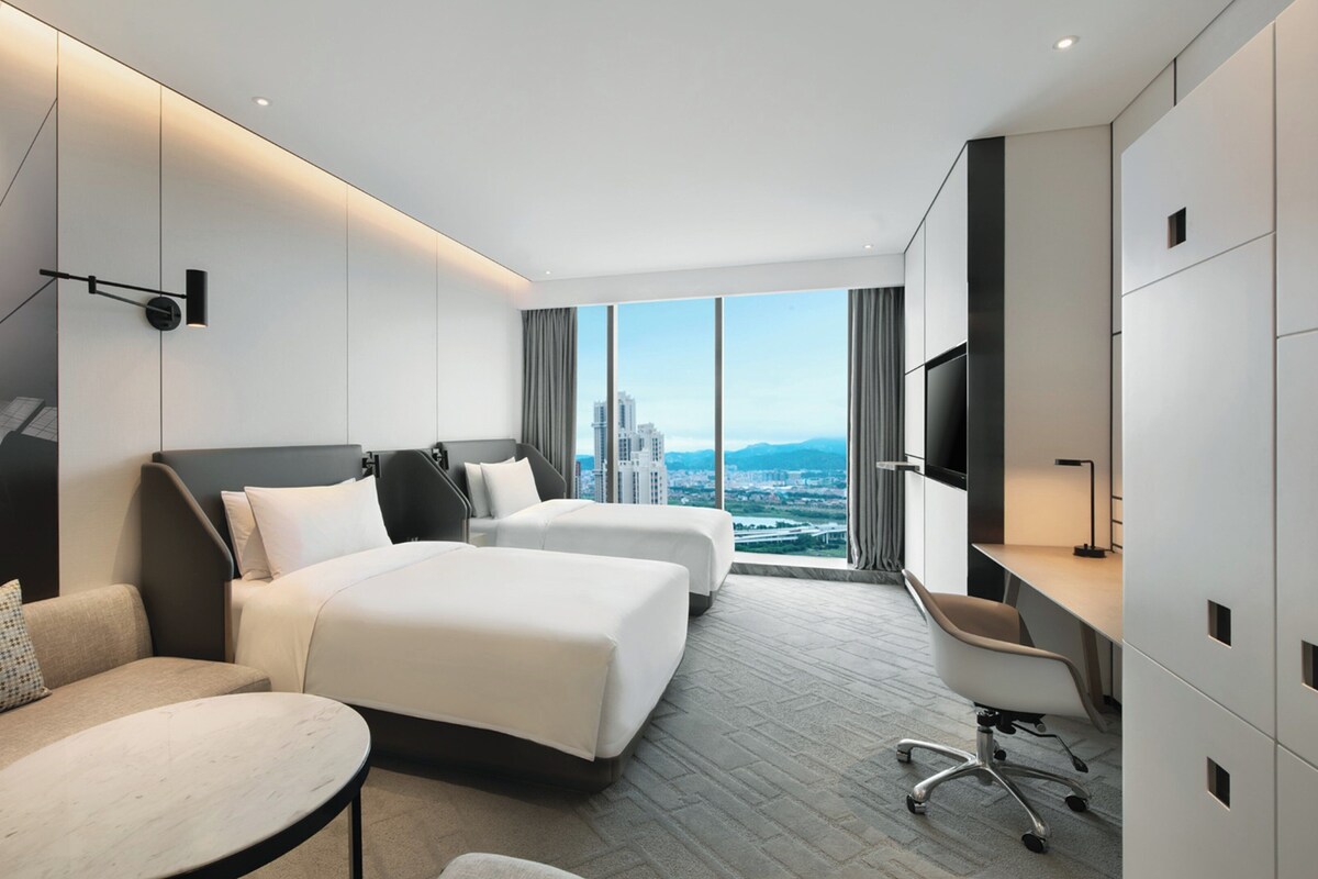 Une chambre d’hôtel moderne équipée de deux lits, d’un bureau, d’une chaise et d’une grande fenêtre donnant sur la ville.
