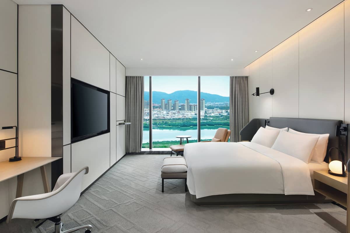 A modern hotel room with a large bed, a desk, a TV, and a view of a cityscape through large windows.