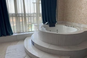 A modern bathroom with a large, circular jacuzzi tub, a window with sheer curtains, and a tiled floor.