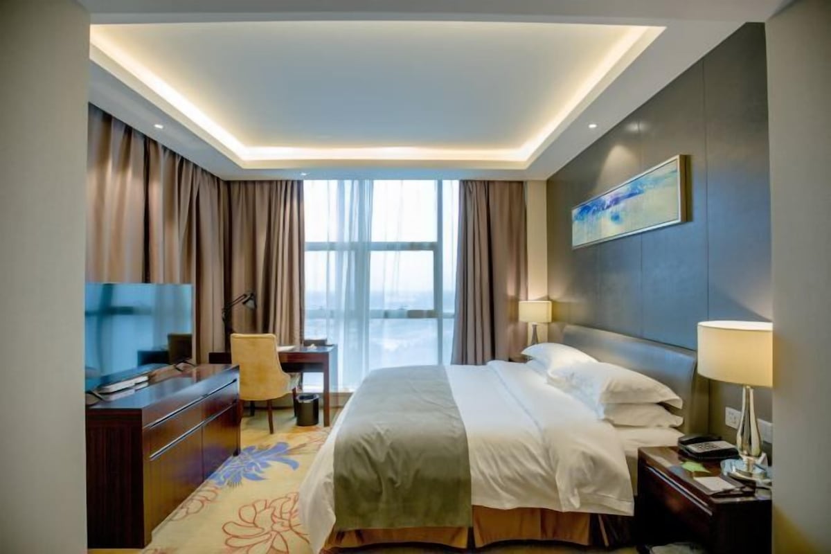 A hotel room with a bed, desk, chair, TV, and a large window.