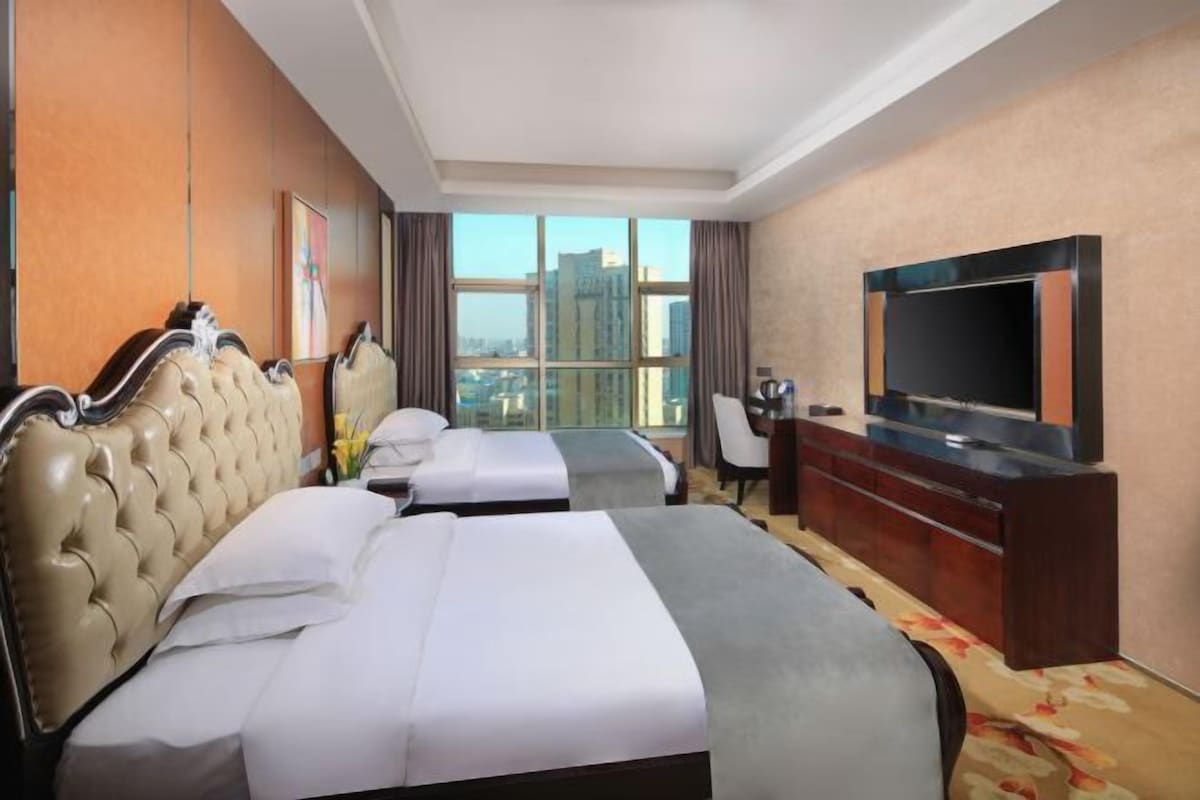 A hotel room with two beds, a TV, a desk, and a large window with a city view.