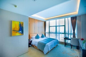 A hotel room with a bed, a desk, a chair, a large window, and a painting on the wall.