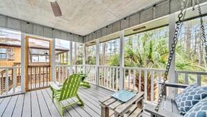 Eine überdachte Veranda mit Holzboden, einem grünen Adirondack-Sessel, einem Holztisch und Blick auf üppige tropische Vegetation.