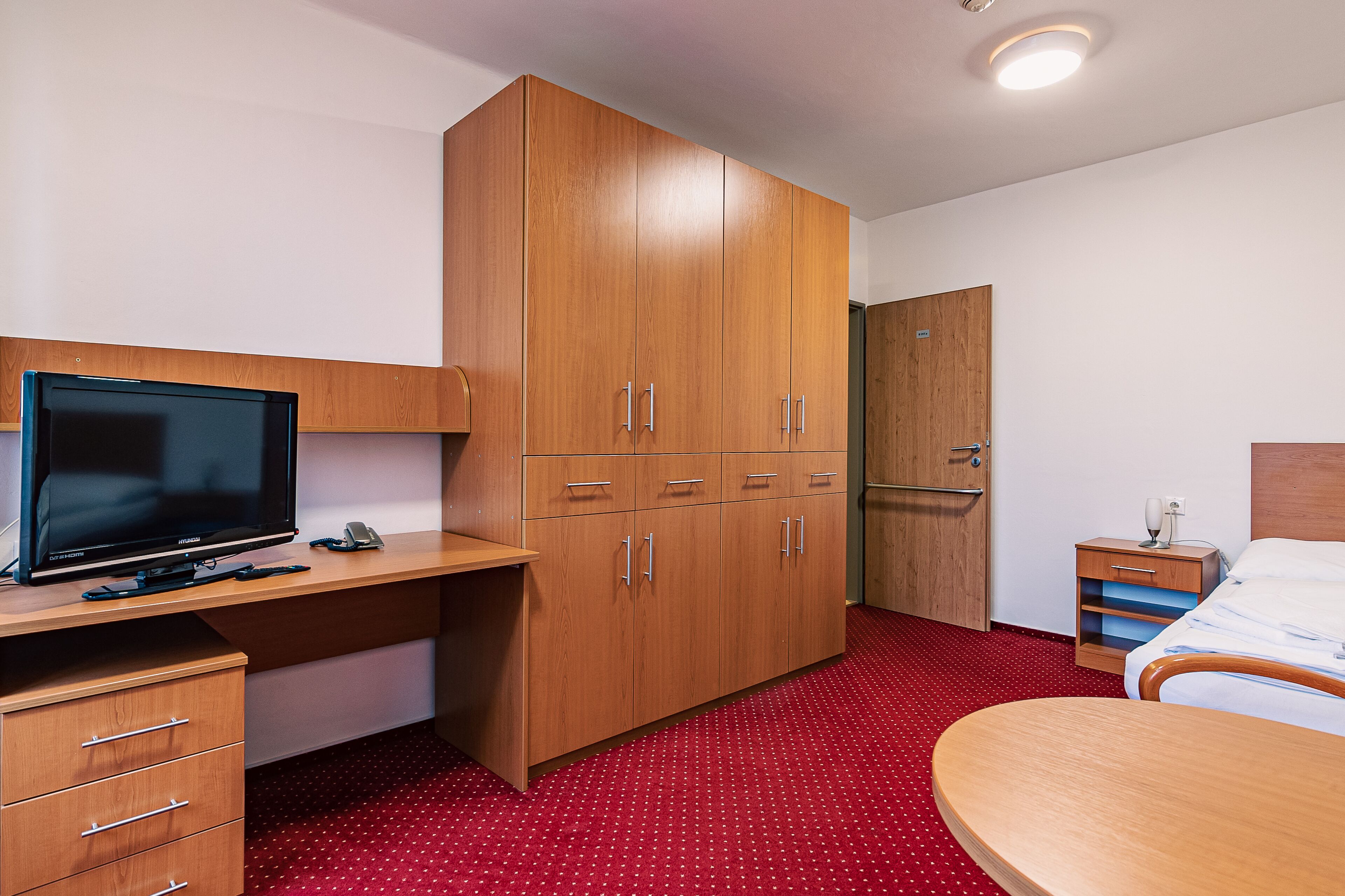 A hotel room with a wooden desk, a television, a wardrobe, a bed, and a nightstand.