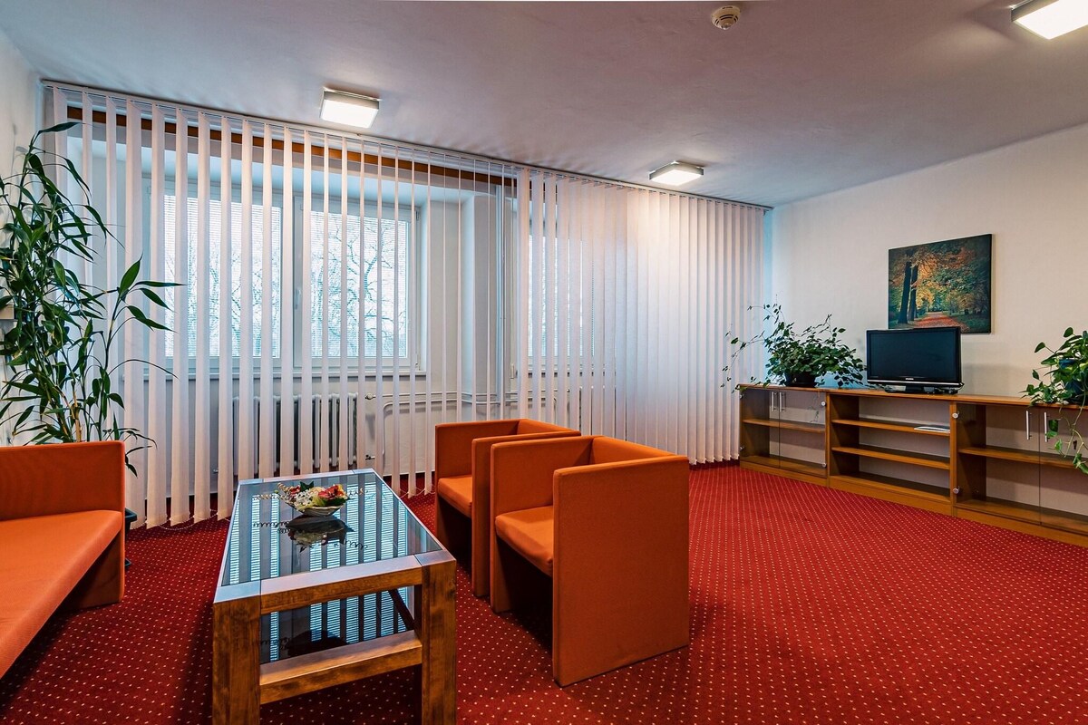 A room with a red carpet, orange seating, a glass-top table, a TV, and a painting on the wall.