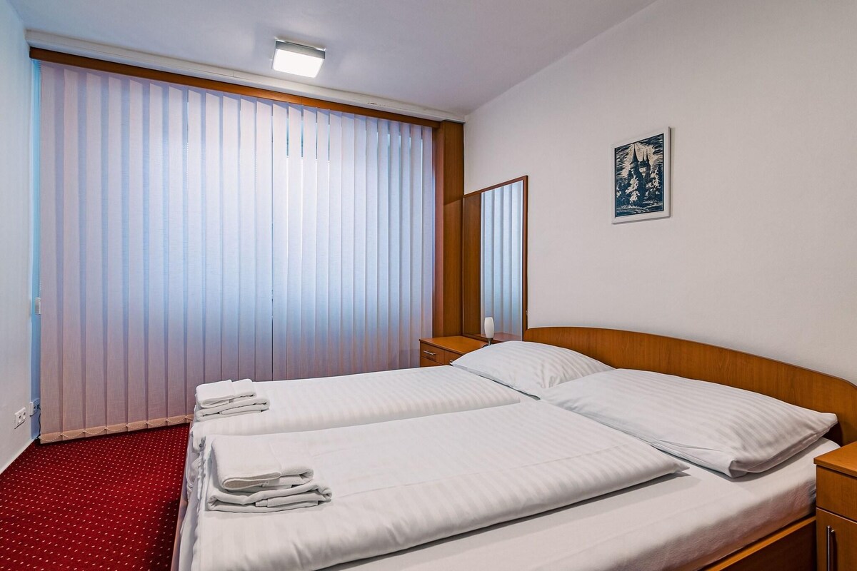 A hotel room with two beds, a red carpet, a wooden headboard, a bedside table, a nightstand, a wall-mounted picture, and a window with vertical blinds.