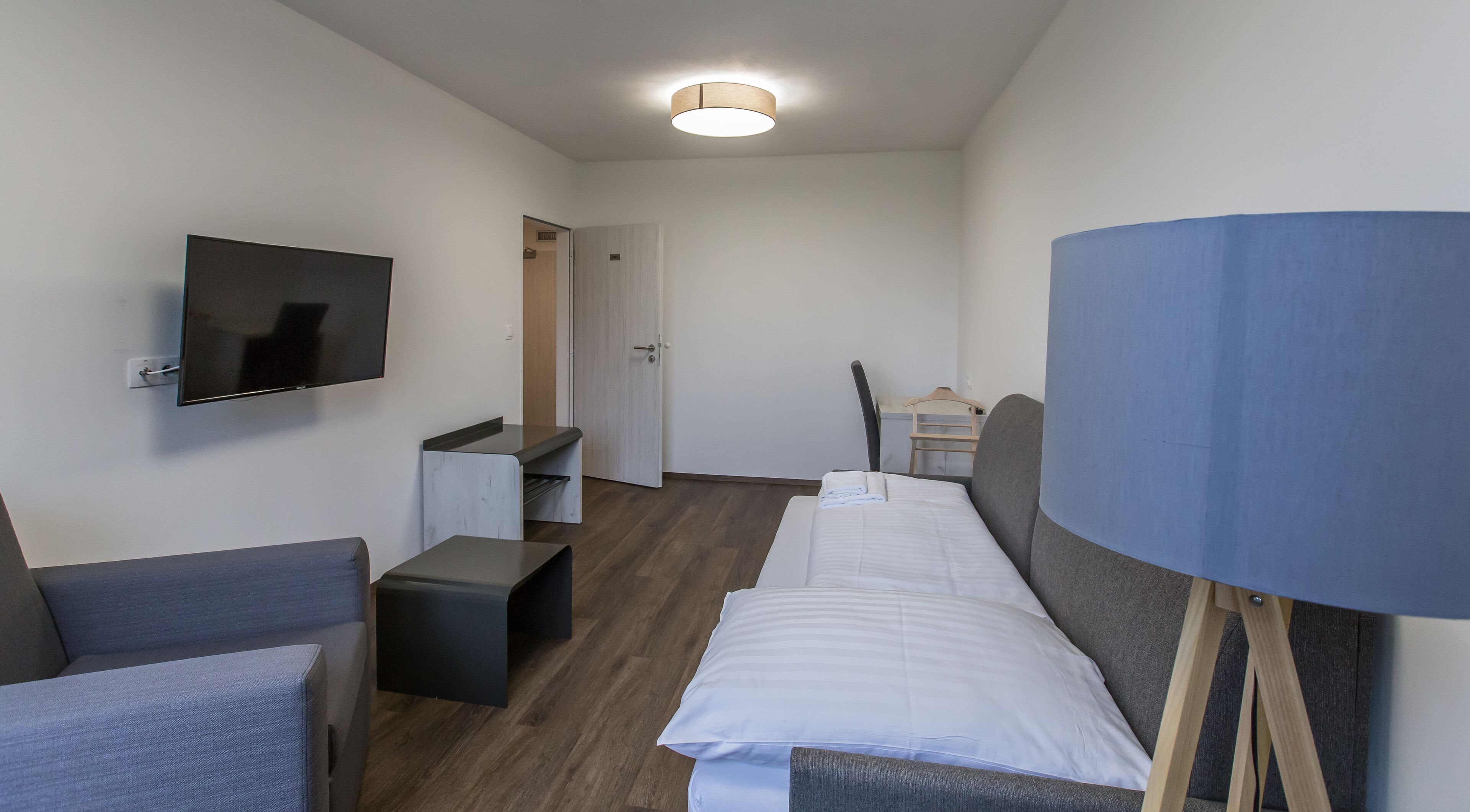 Quarto de hotel com uma cama, televisão na parede, escrivaninha e uma cadeira.