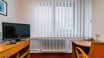 A hotel room with a television on a wooden desk, a radiator, and a window with blinds.