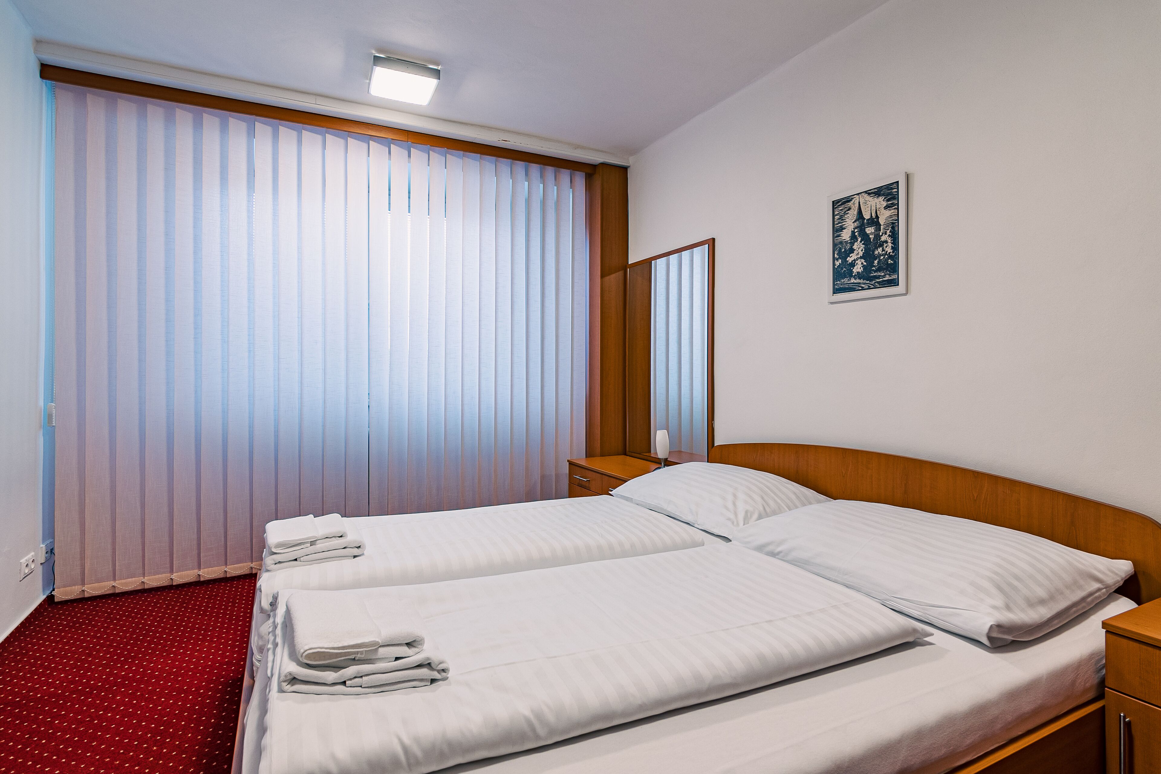 A hotel room with two beds, a wooden headboard, a bedside table, a nightstand, a red carpet, and a large window with vertical blinds.