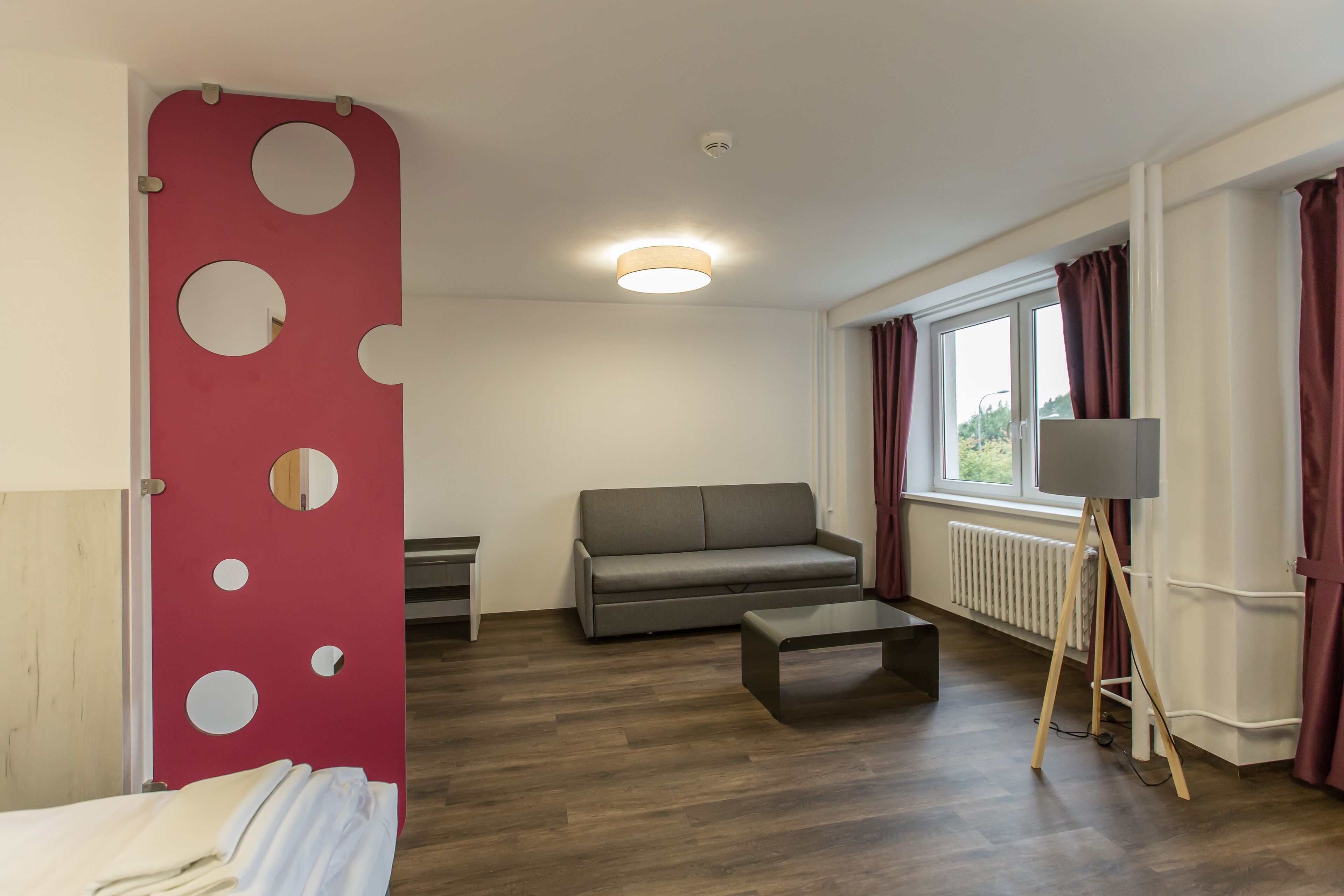 A modern room with a grey sofa, a small table, a red accent wall with circular cutouts, a standing lamp, and a window with curtains.