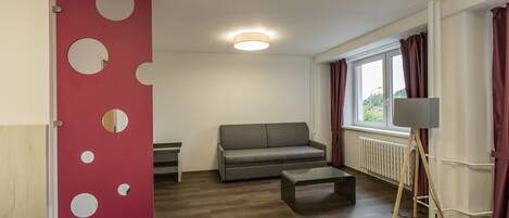 A modern room with a grey sofa, a small table, a red accent wall with circular cutouts, a standing lamp, and a window with curtains.