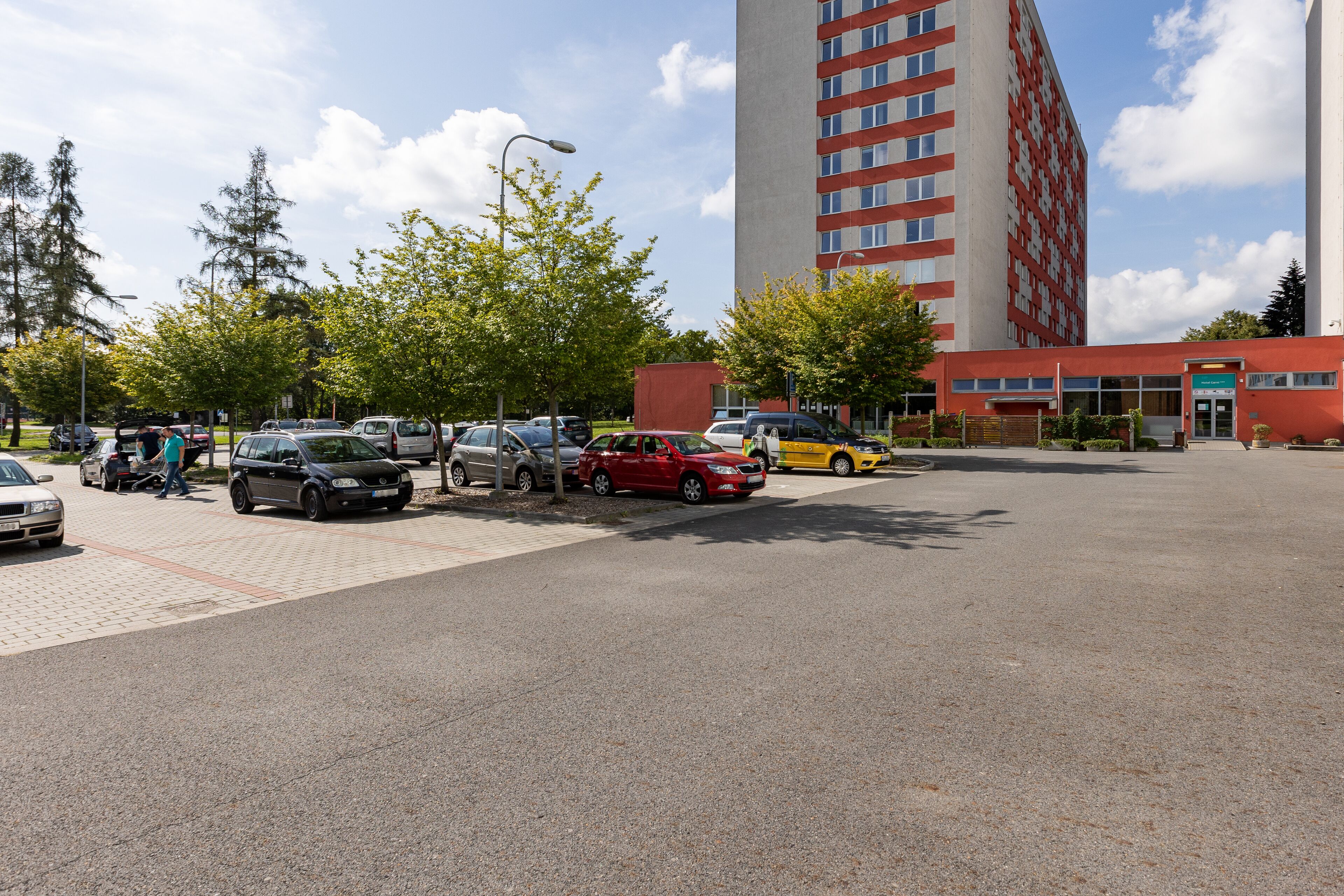 Un parking avec plusieurs voitures, un bâtiment à la façade rouge et des arbres en arrière-plan.
