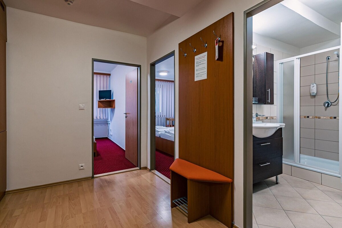 A hotel room with a wooden bench, a bathroom with a shower, and a hallway leading to a bedroom with a TV.