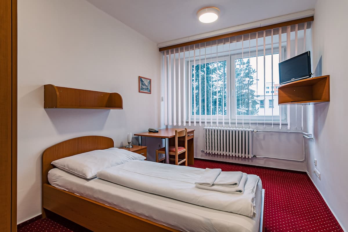 A hotel room with a bed, a desk, a chair, a TV on a shelf, and a window with blinds.