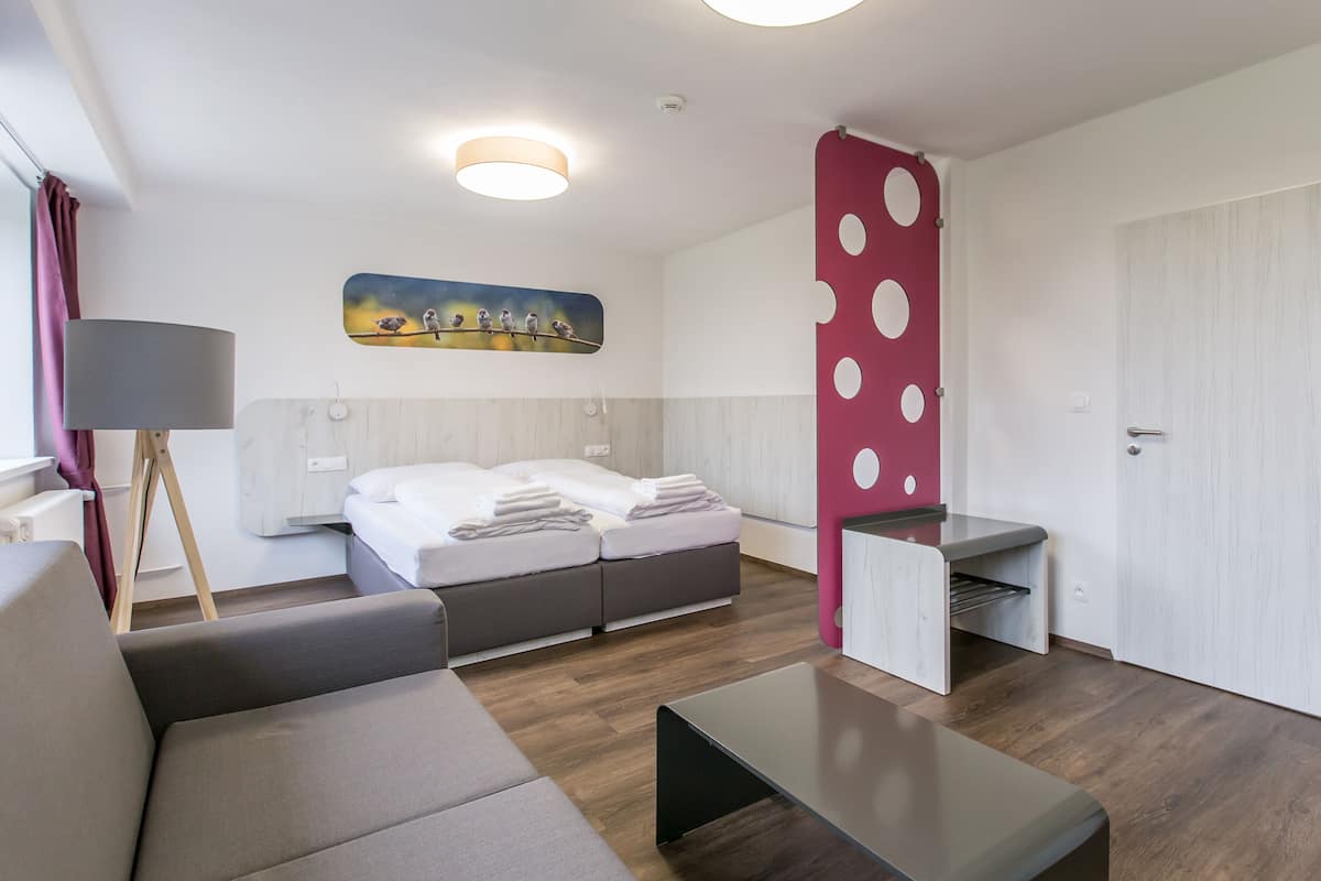 A modern hotel room with a bed, a sofa, a desk, and a painting of birds on the wall.