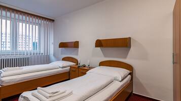 A hotel room with two single beds, a wooden nightstand, a small table, and a window with blinds.