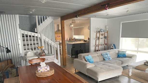A spacious living area with a wooden dining table, a sofa with decorative pillows, a kitchen in the background, and a staircase leading to an upper level.