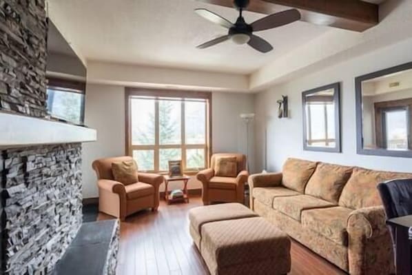 A living room with a stone fireplace, two armchairs, a sofa, and a ceiling fan. - Sable Ridge View Suite - Fireplace - Pool - A/C. (Radium Hot Springs)
