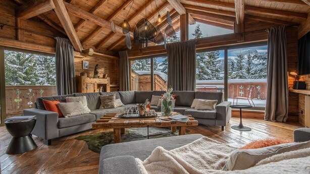 A cozy log cabin interior with a wooden ceiling, a large sofa set, a coffee table, and a view of snowy mountains through large windows. - Luxury Chalet in Courchevel with Pool, Hammam and Sauna (Courchevel 1850)