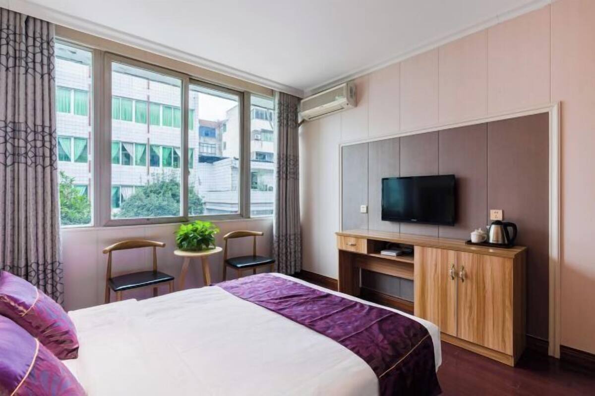 Habitación de hotel con una cama grande, televisión, una pequeña mesa con sillas y ventana con vistas a edificios.