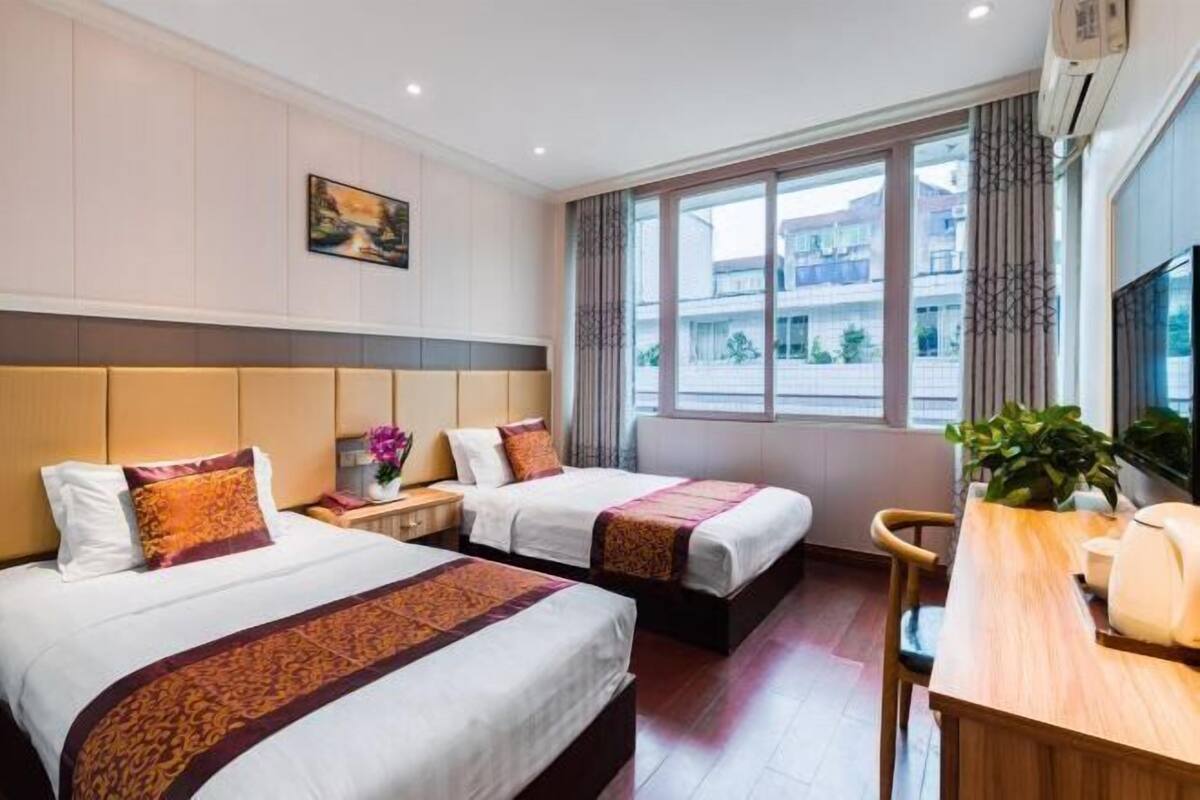 Habitación de hotel con dos camas, un escritorio, una silla, una ventana con vistas a edificios y un cuadro en la pared.