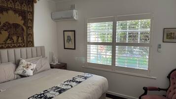 A bedroom with a bed, a nightstand, a chair, a window with blinds, and an air conditioning unit.