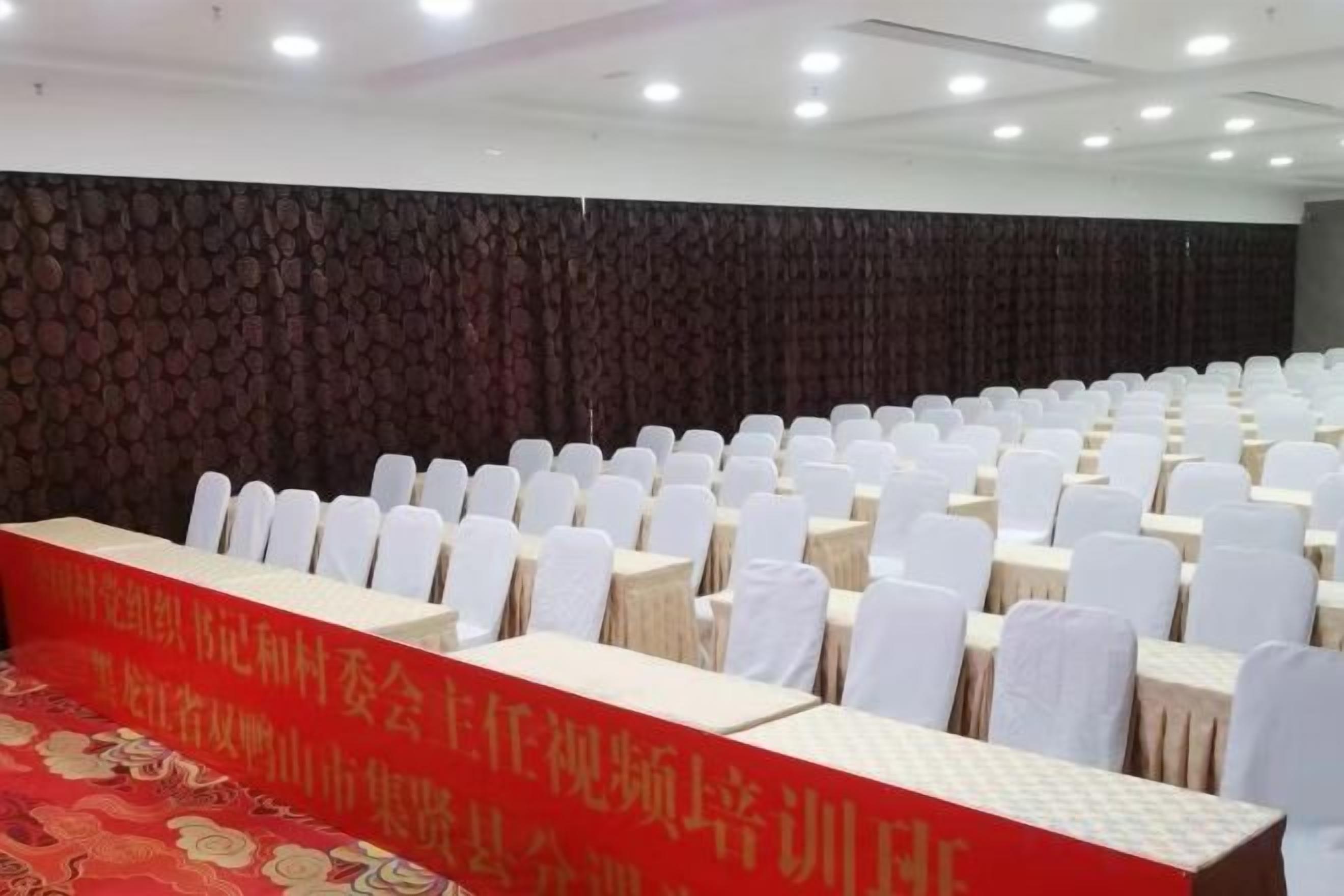 A conference room with rows of white chairs and tables, a red banner with text, and a patterned wall.