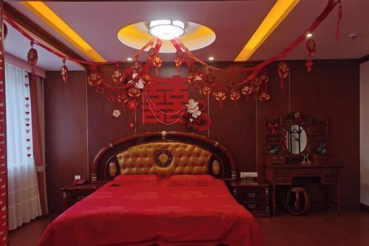 A bedroom with a large bed, wooden headboard, and red bedding. There is a dresser with a mirror and a small table beside the bed. The room has a warm color scheme with red and brown tones.