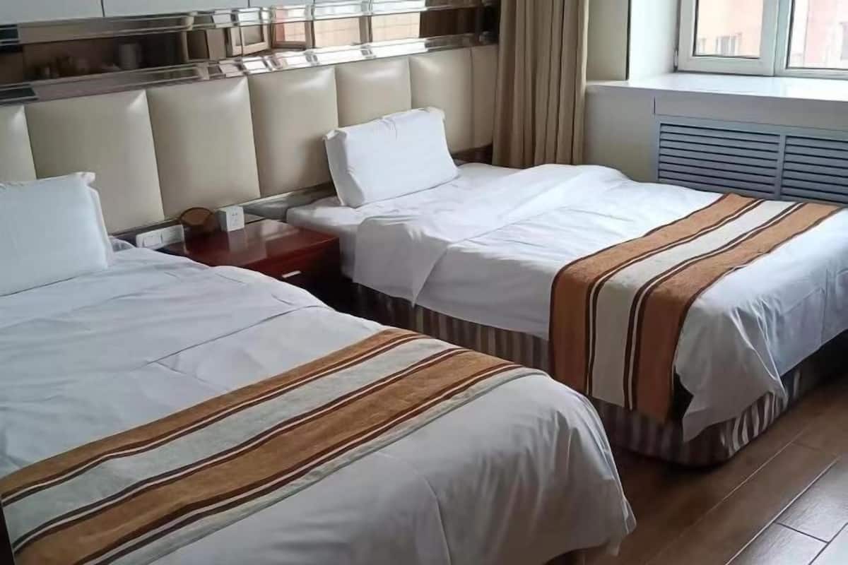 A hotel room with two beds, a nightstand, a window with curtains, and a radiator.