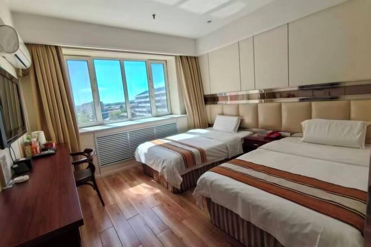 A hotel room with two beds, a desk, a chair, a window with curtains, and a view of the city.