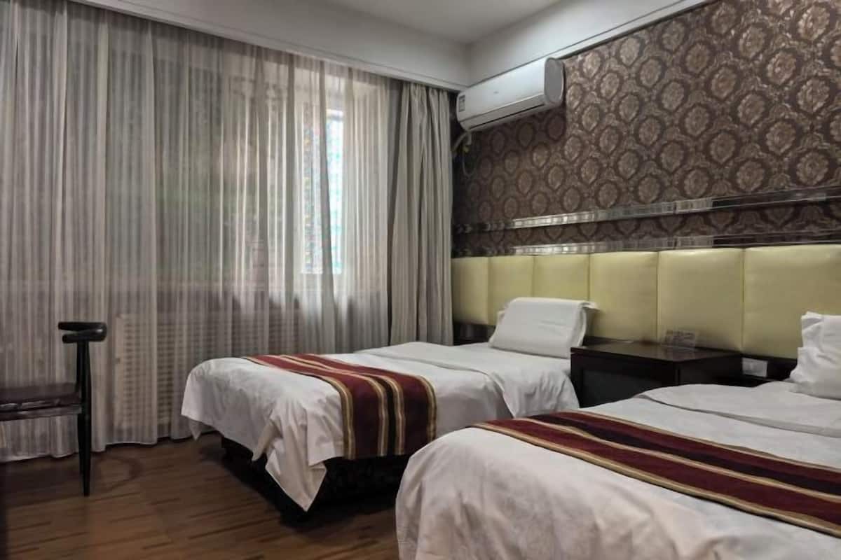 A hotel room with two beds, a window with curtains, an air conditioning unit, and a patterned wall.