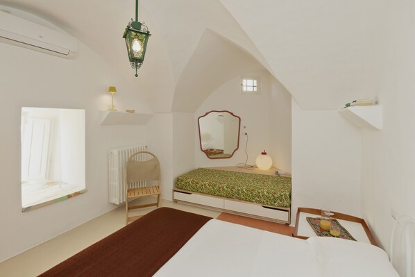 A bedroom with a sloped ceiling, a bed with a floral bedspread, a bedside table with a lamp, a window, and a radiator.