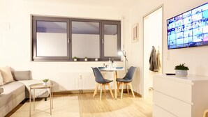 Ein modernes Wohnzimmer mit einer Couch, einem kleinen Tisch mit einer Pflanze, einem Essbereich mit Stühlen, einem an der Wand montierten Fernseher und einer weißen Kommode.