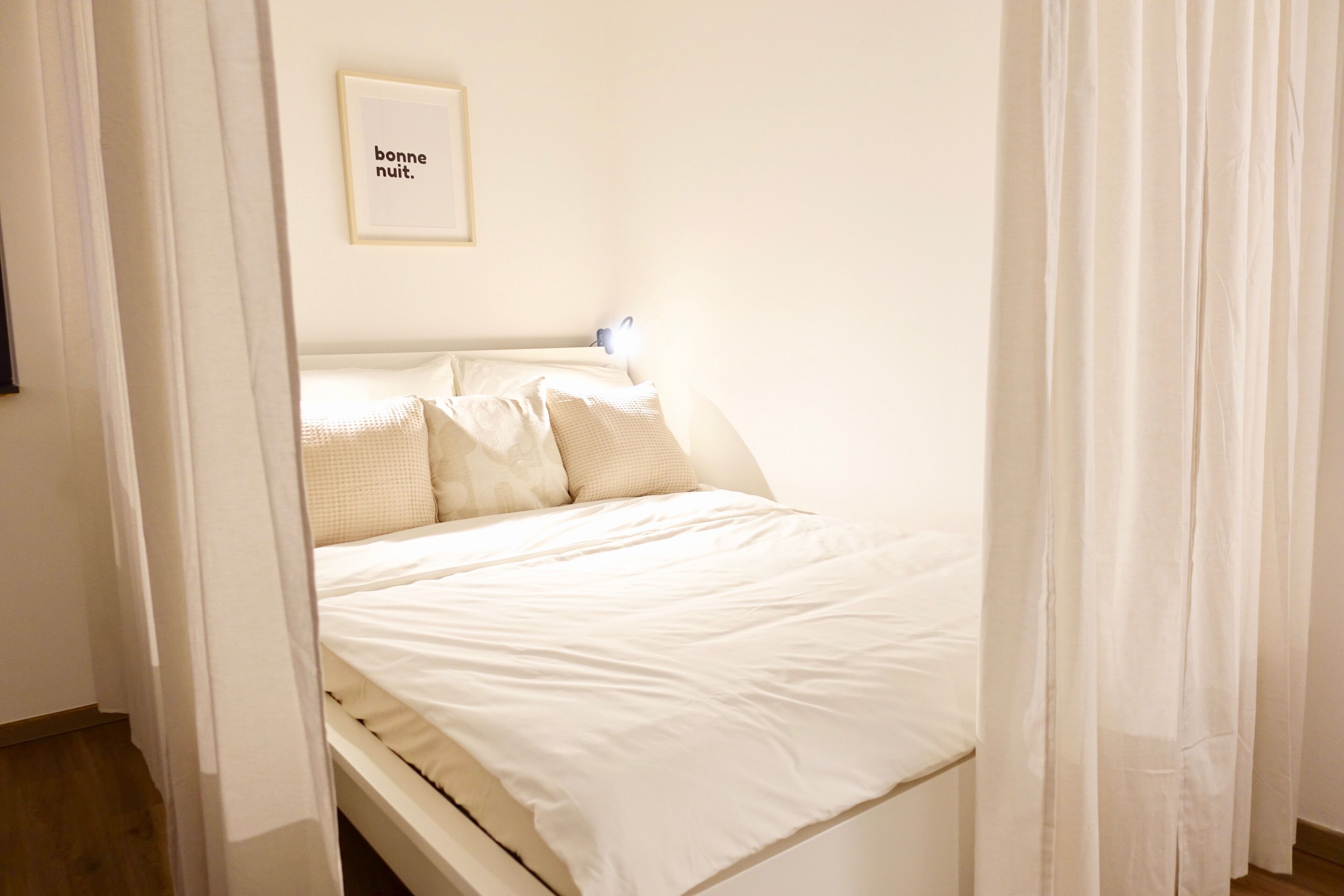 A neatly made bed with white bedding and pillows, a bedside table with a lamp, and a framed sign on the wall.