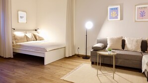 Ein Schlafzimmer mit einem Bett, einem Nachttisch, einer Stehlampe, einem Sofa und einem kleinen Tisch mit einer Pflanze. An der Wand hängen zwei gerahmte Bilder.