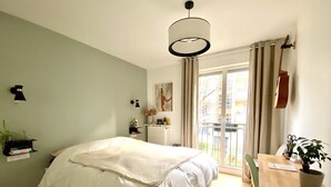 Un dormitorio con cama, escritorio, silla, radiador, ventana con cortinas y una lámpara colgante.