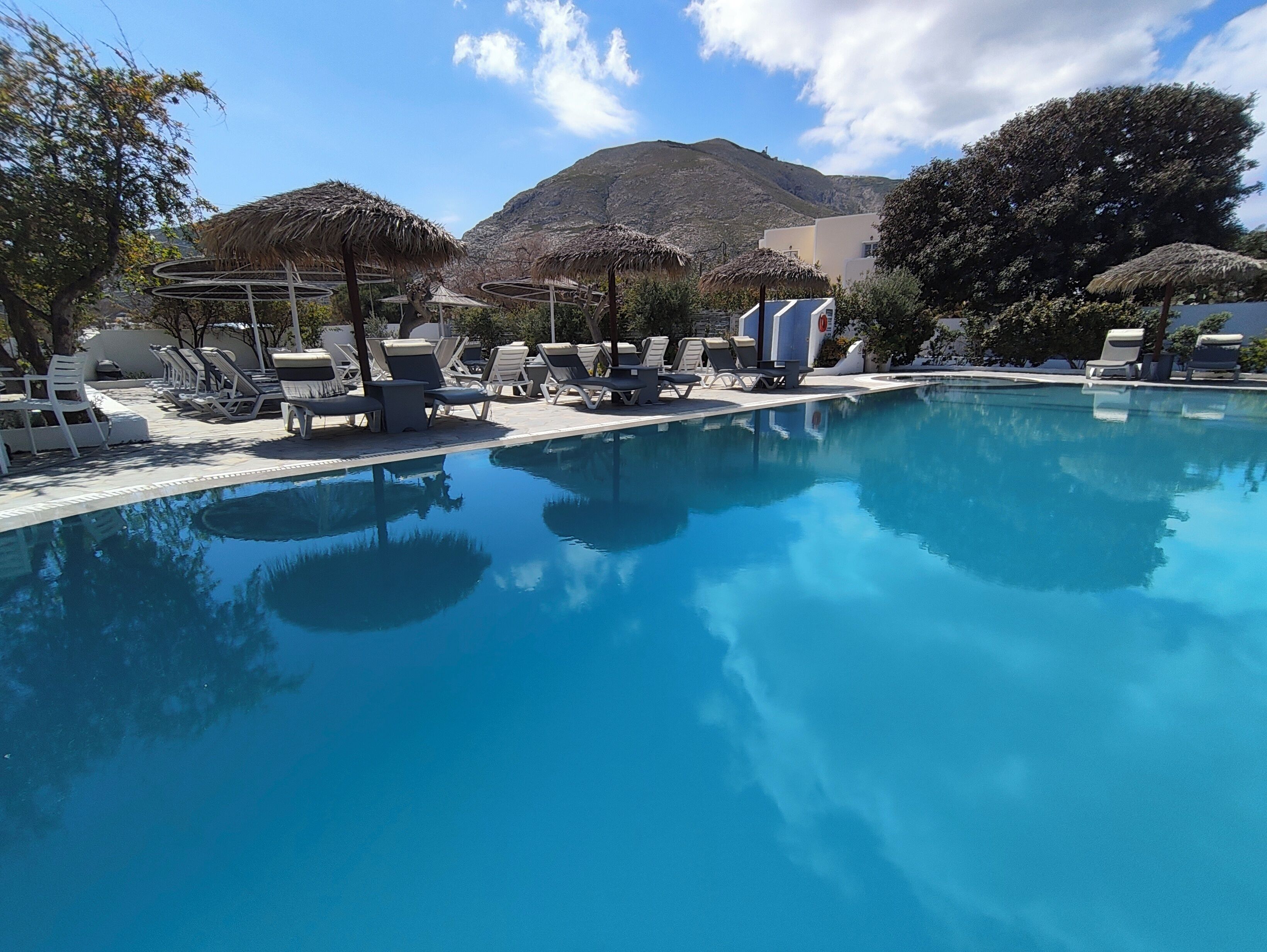 Una piscina con sillas de descanso y sombrillas de paja, rodeada de árboles y una montaña al fondo.
