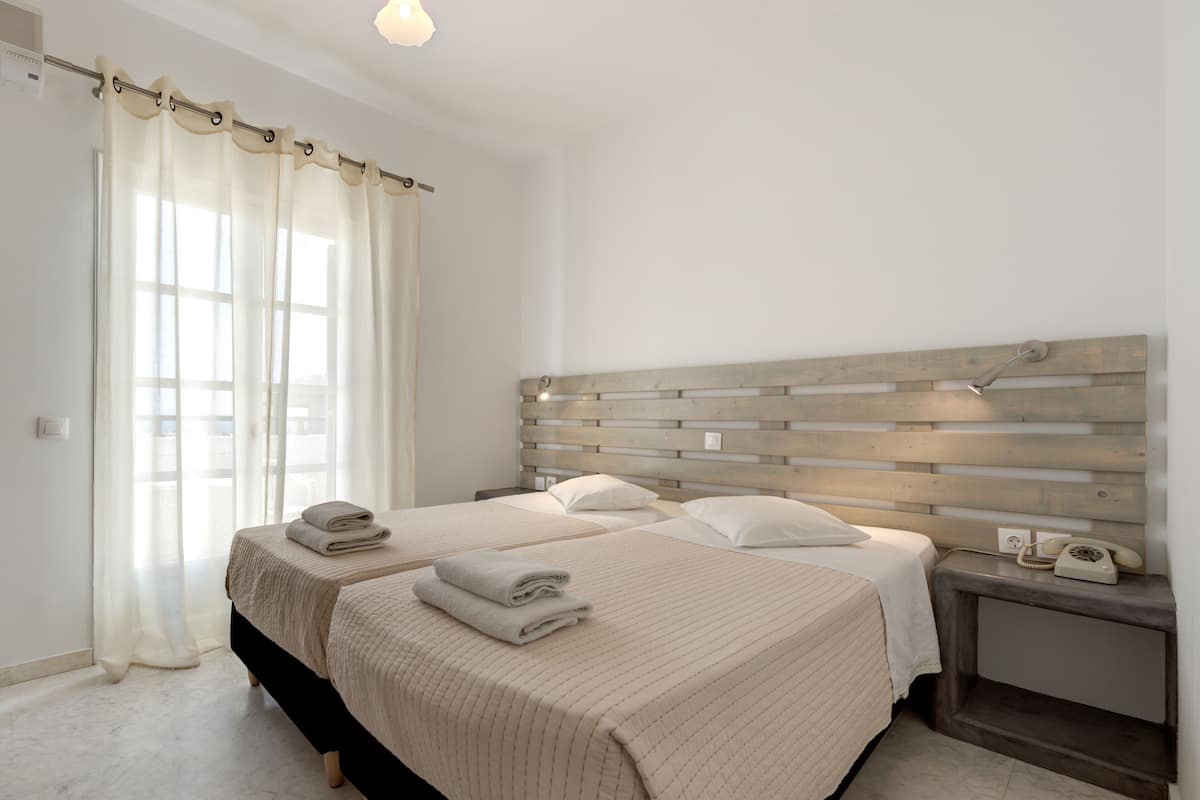 Une chambre d’hôtel avec deux lits, une tête de lit en bois, une table de chevet avec un téléphone et une fenêtre avec des rideaux légers.