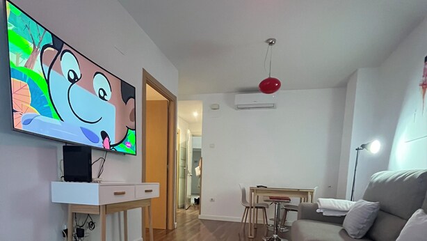 A modern living room with a flat-screen TV, a grey sofa, a dining area, and a red pendant light.