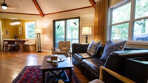 A cozy living room with a wooden floor, a leather sofa, a dining table, and a large window with curtains. - Unique Cabin w/ Riverside View + Hot Tub + Apple Pie (Ellijay)