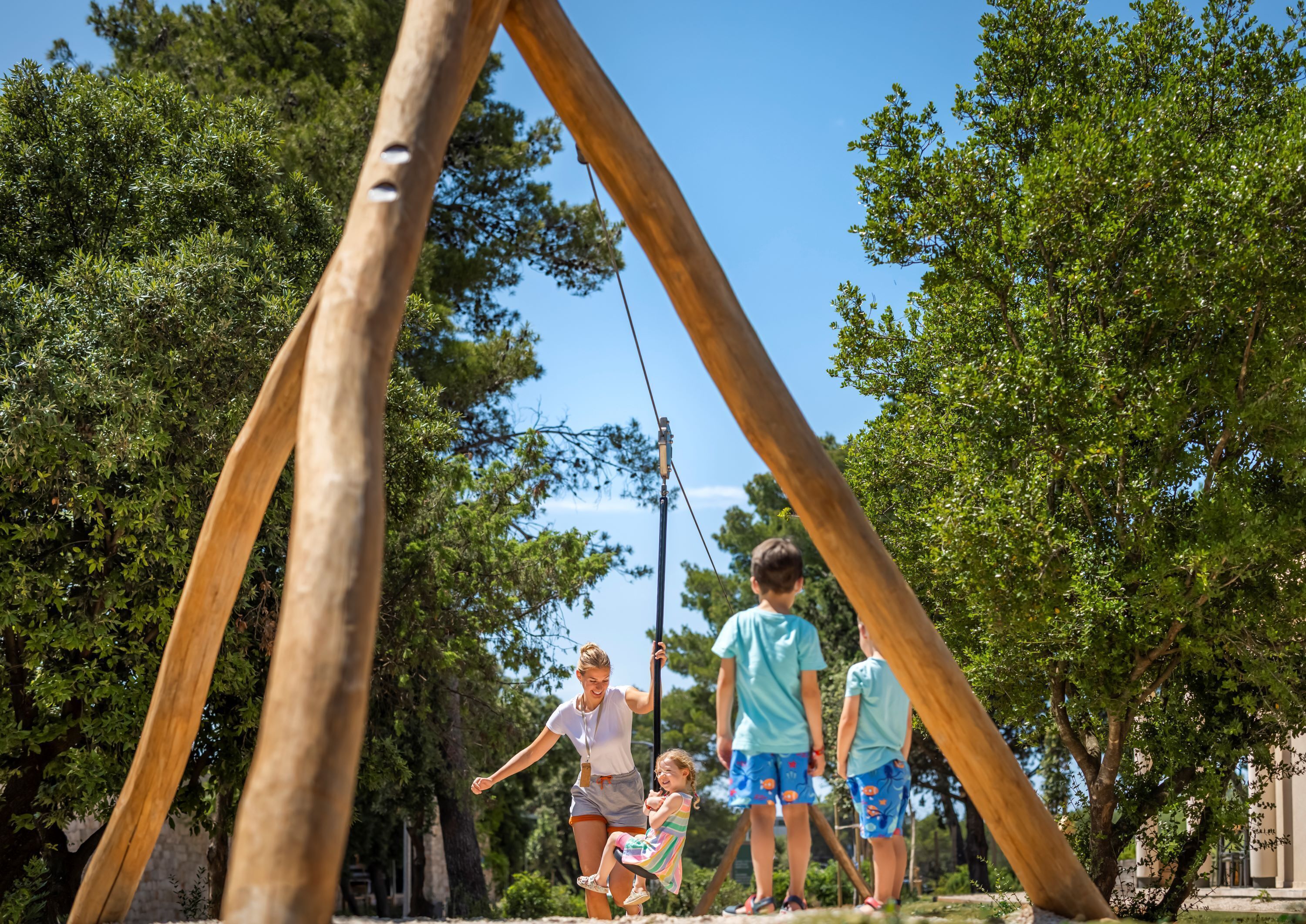 Ein hölzernes Schaukelgerüst in einem Park mit Bäumen und blauem Himmel.