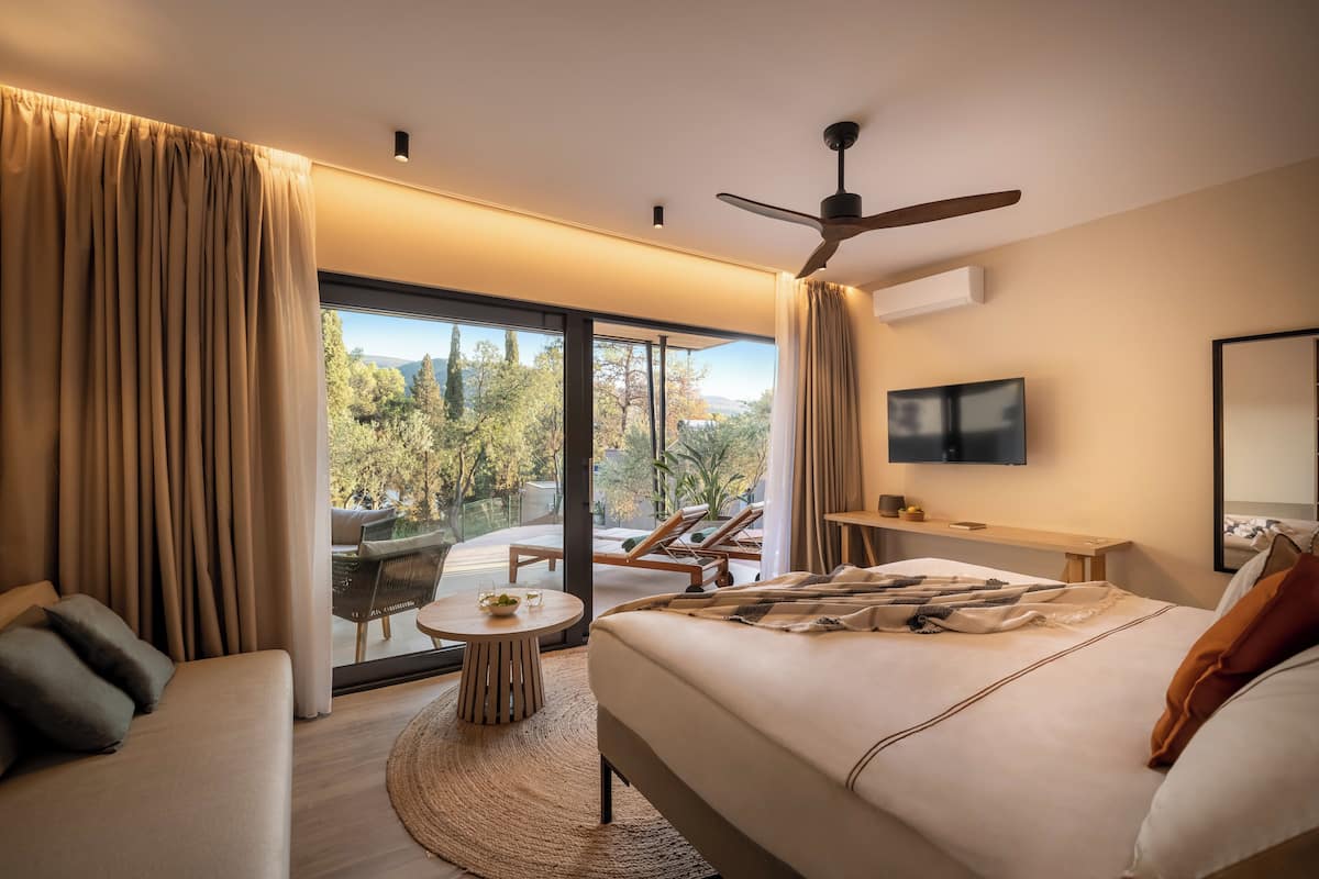 Een moderne hotelkamer met een groot bed, een bureau, een flatscreen televisie en een balkon met uitzicht op bomen en een stoel.