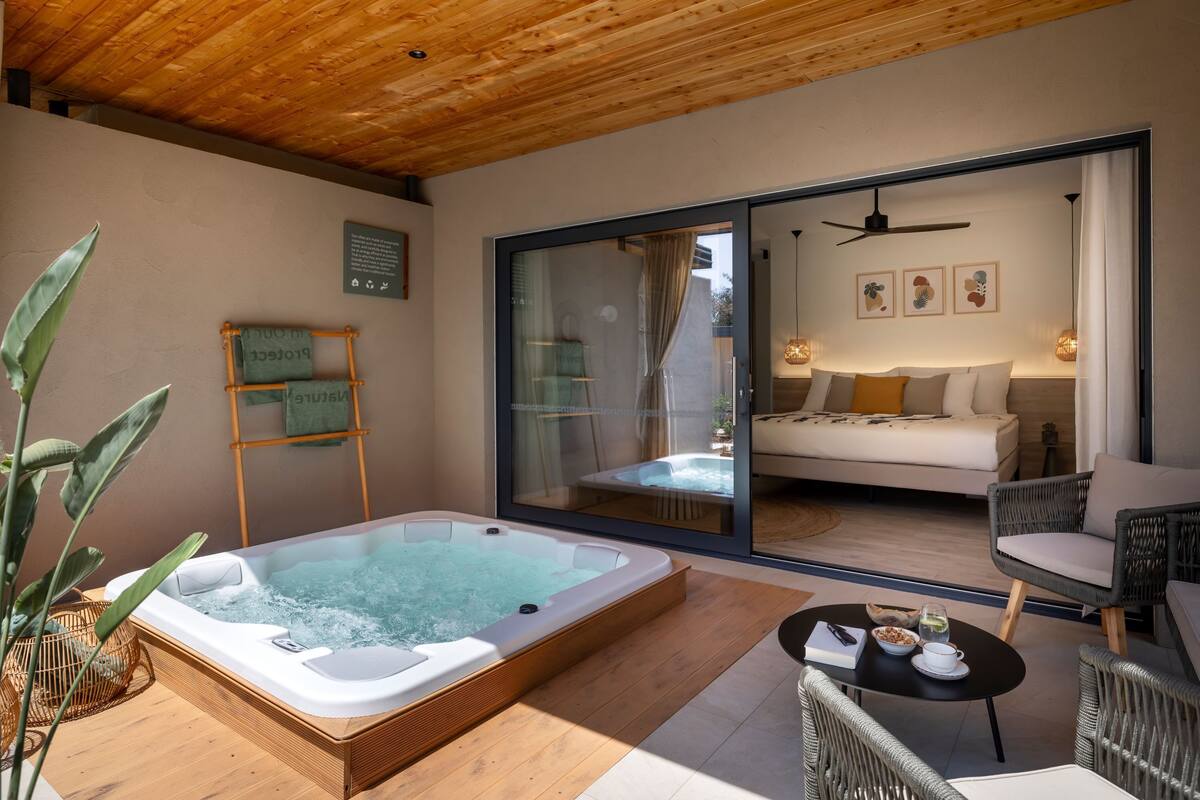 Een moderne spa met een bubbelbad, rieten meubels en een glazen schuifdeur naar een slaapkamer met een bed en een slaapbank.