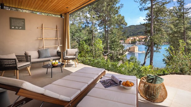 A modern outdoor seating area with a sofa, chairs, and a lounge chair, overlooking a scenic coastal view with trees and a distant town.