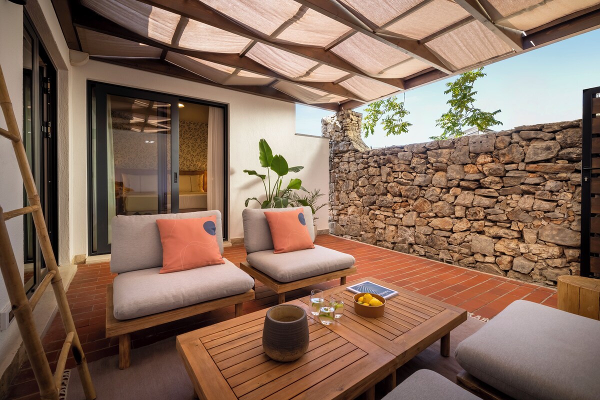 A covered outdoor seating area with two cushioned chairs, a wooden table, and a stone wall.