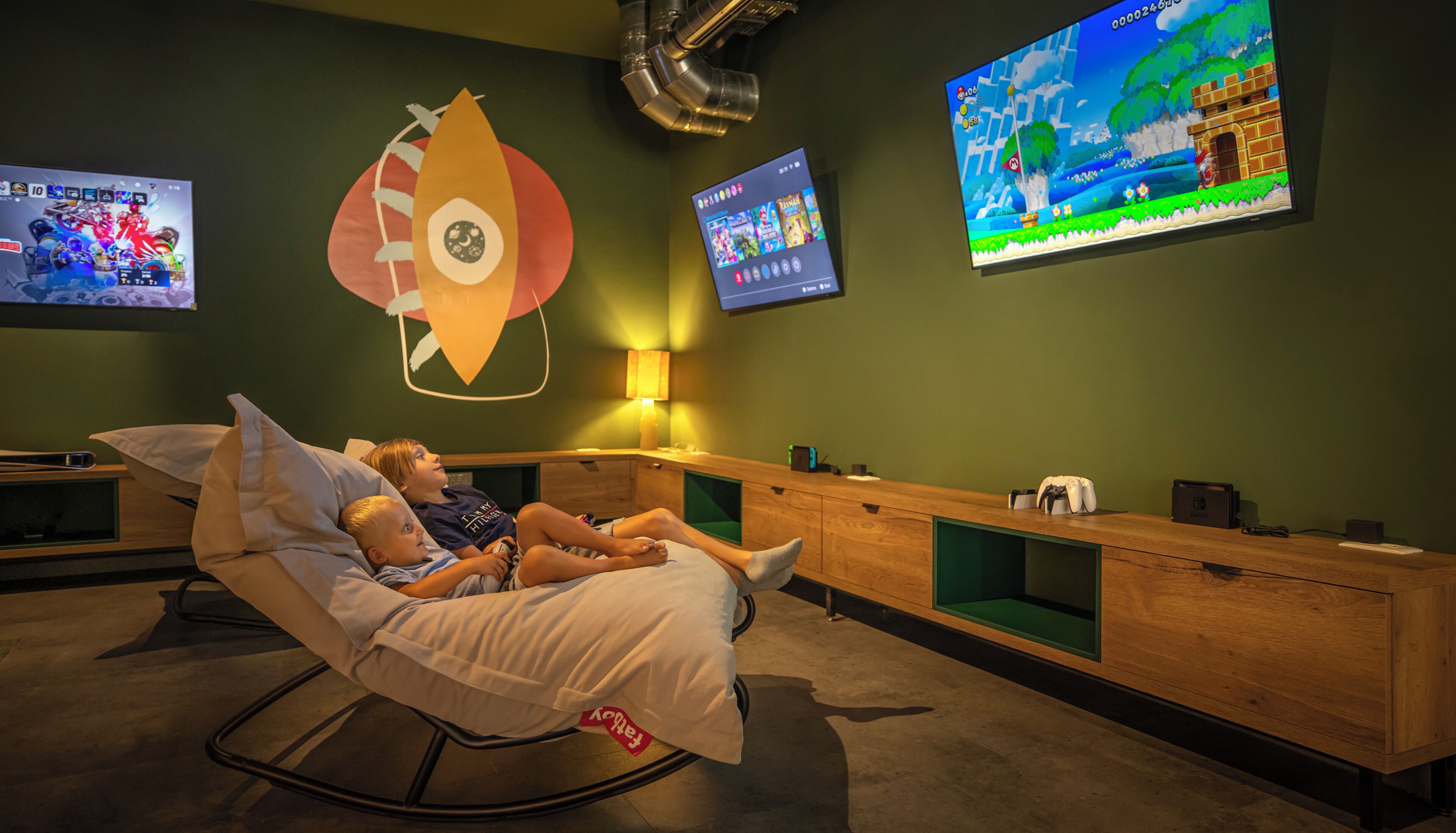 Une pièce comprenant un fauteuil inclinable, un meuble en bois et deux écrans diffusant des jeux vidéo.