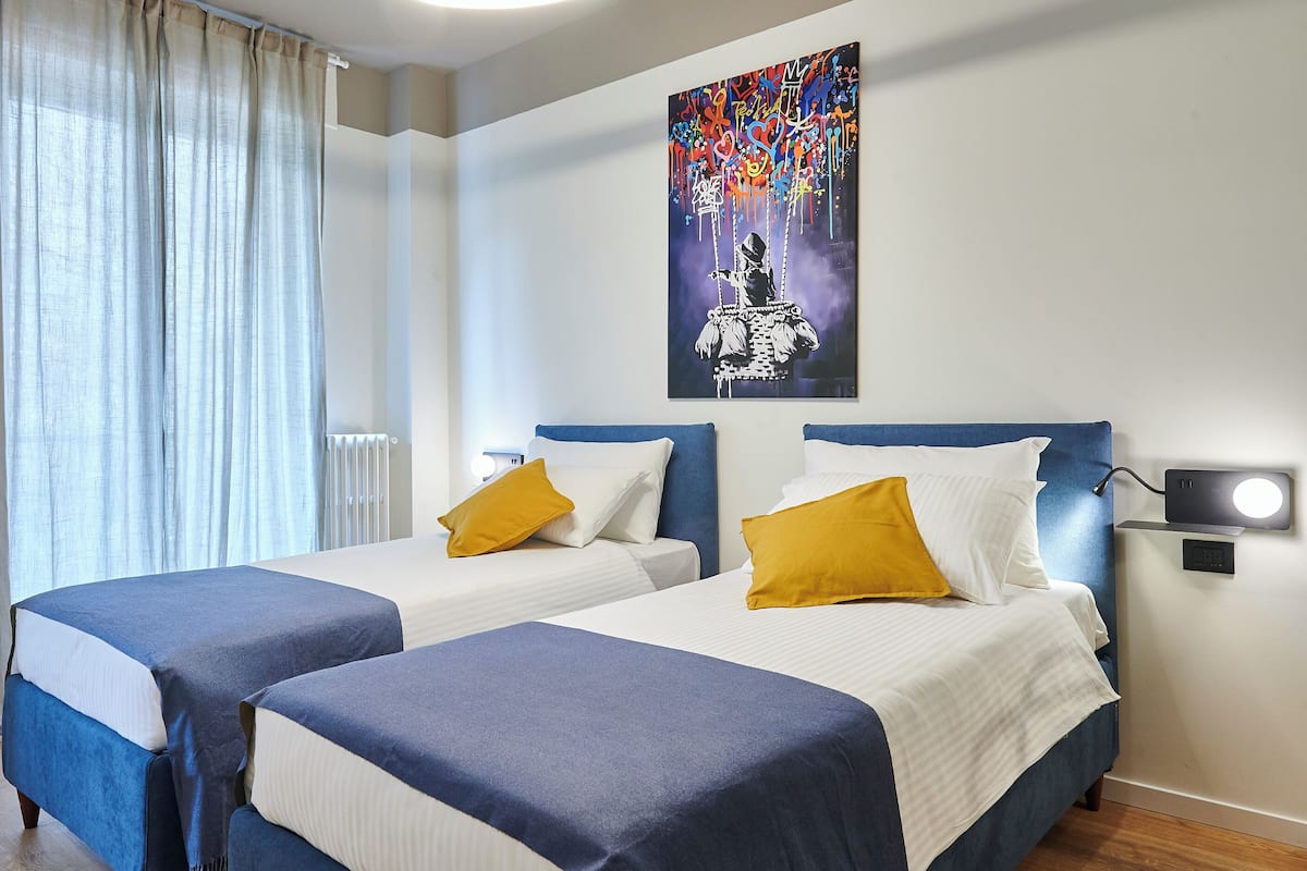 A hotel room with two beds, a nightstand with a lamp, a painting on the wall, and a window with curtains.