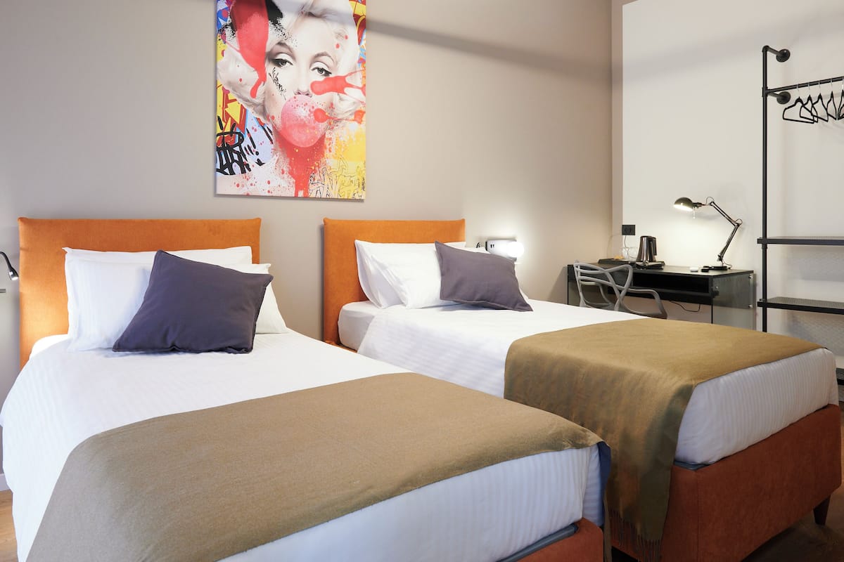 A hotel room with two beds, each with a headboard, white linens, and a brown throw blanket. There is a desk with a lamp, a chair, and a coat rack. A colorful artwork is on the wall above the beds.
