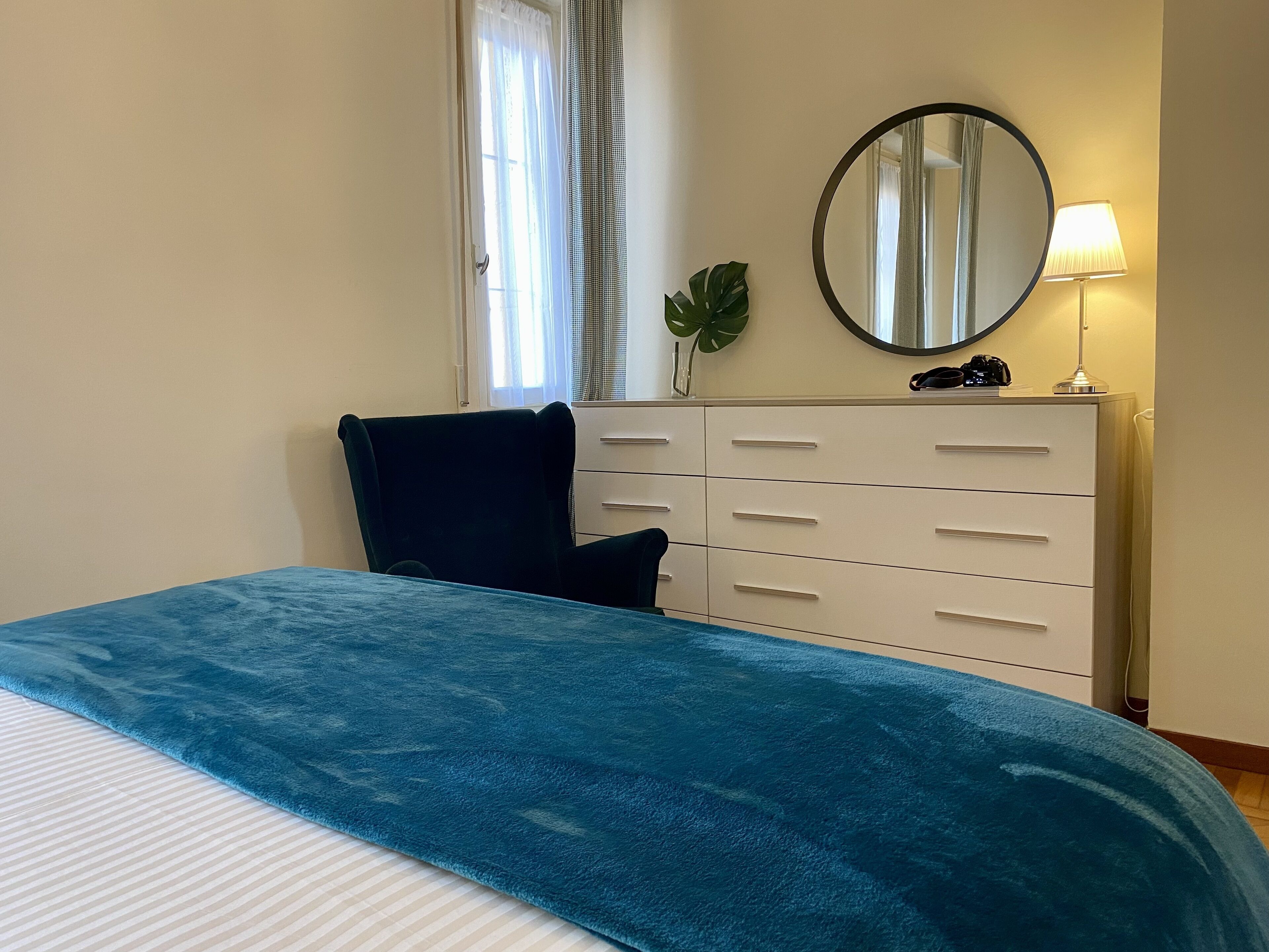 A bedroom with a bed, a dresser, a mirror, a lamp, and a window with curtains.