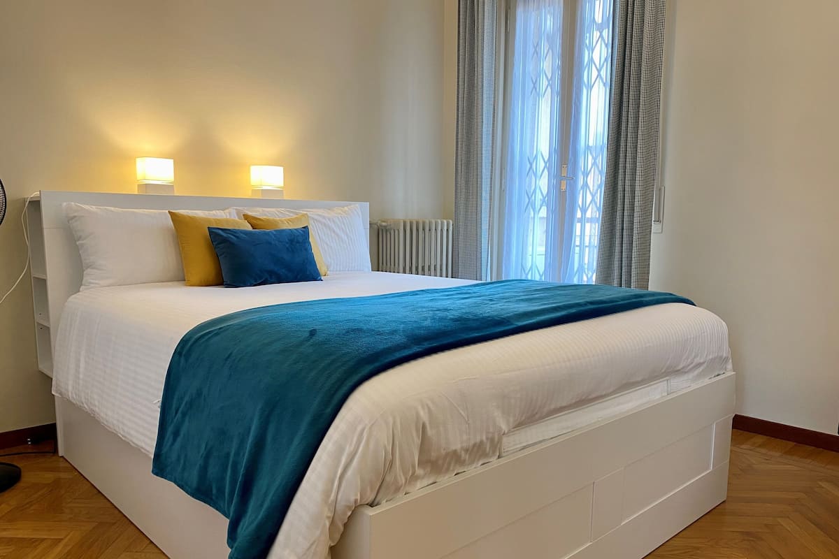 A neatly made bed with a white headboard, blue and yellow pillows, and a blue blanket. There are two bedside lamps and a window with curtains.